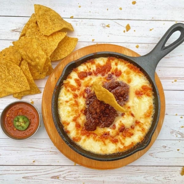 Melted cheese with chorizo in a skillet, served with tortilla chips and salsa on a white wooden table.