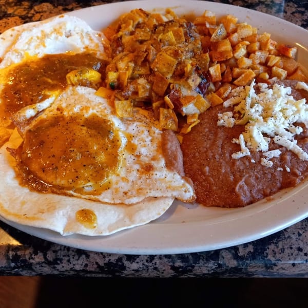 Huevos Rancheros with two fried eggs, salsa, refried beans, and diced potatoes on a white plate.