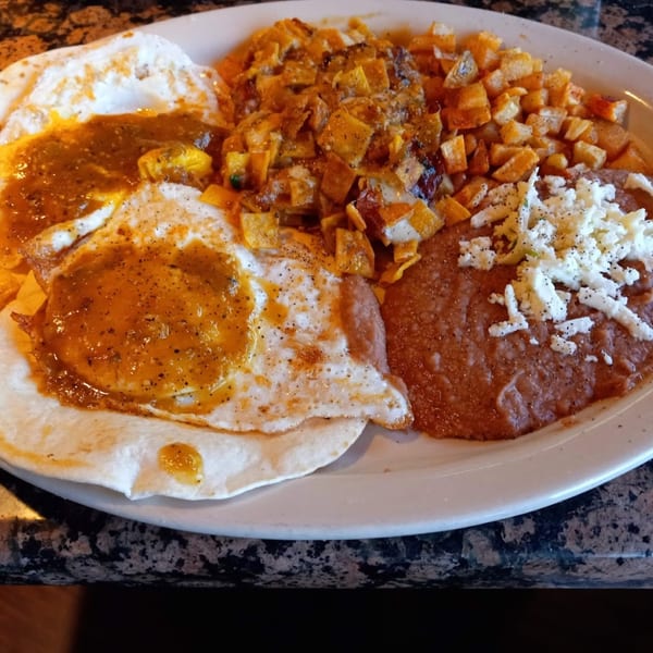 Huevos rancheros with salsa, refried beans topped with cheese, and diced potatoes on a white plate.