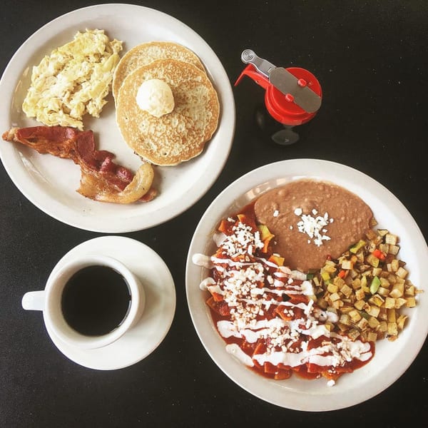Mexican breakfast with chilaquiles, refried beans, potatoes, scrambled eggs, pancakes, bacon, and coffee.